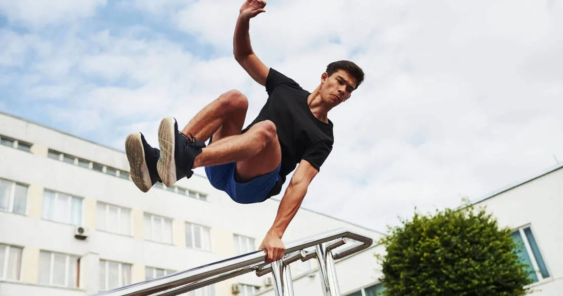 a young man in a black t-shirt and blue shorts is captured mid-air while performing a parkour move, gripping a metal railing…