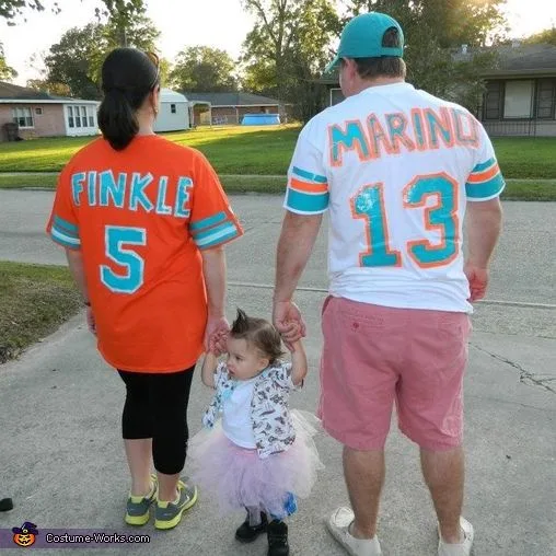 A family of three walks away from the camera down a residential sidewalk. The adult woman wears an orange jersey with…
