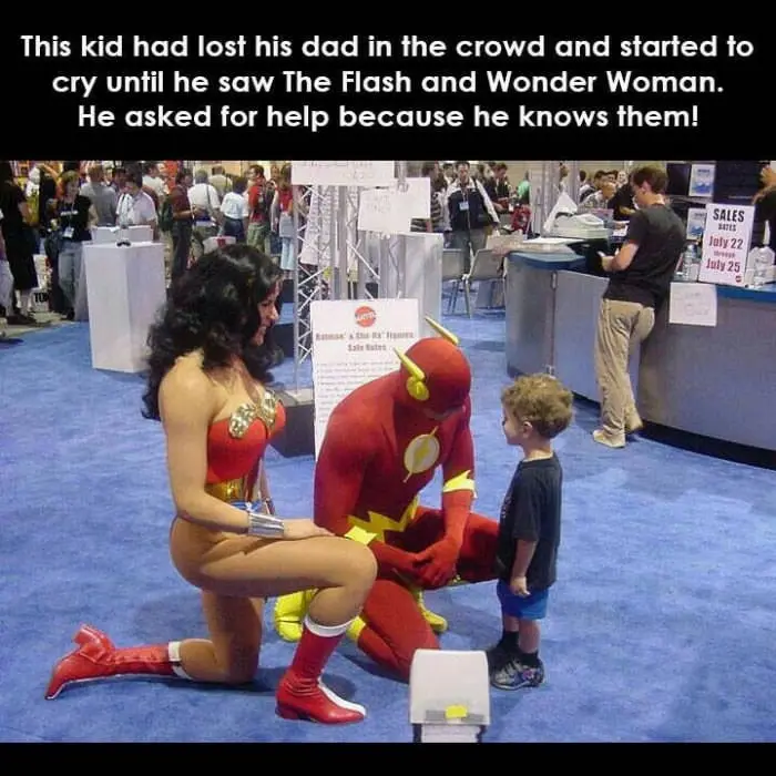 Two adults dressed as The Flash and Wonder Woman are kneeling on a blue carpeted floor at a convention, facing a small child…