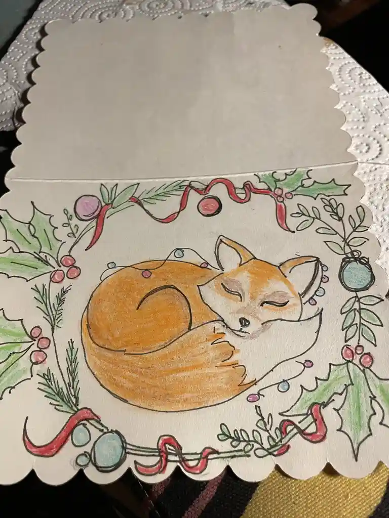 A hand-drawn Christmas card features a curled-up, sleeping orange fox surrounded by a festive wreath of holly, red ribbon…