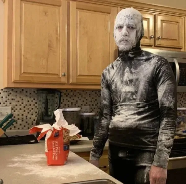 A man stands in a kitchen, completely covered in white flour from head to toe, looking exhausted and defeated. An open bag…