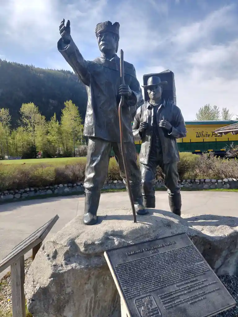 Two bronze statues of Klondike Gold Rush prospectors stand on a large rock, one gesturing upward while holding a staff, the…
