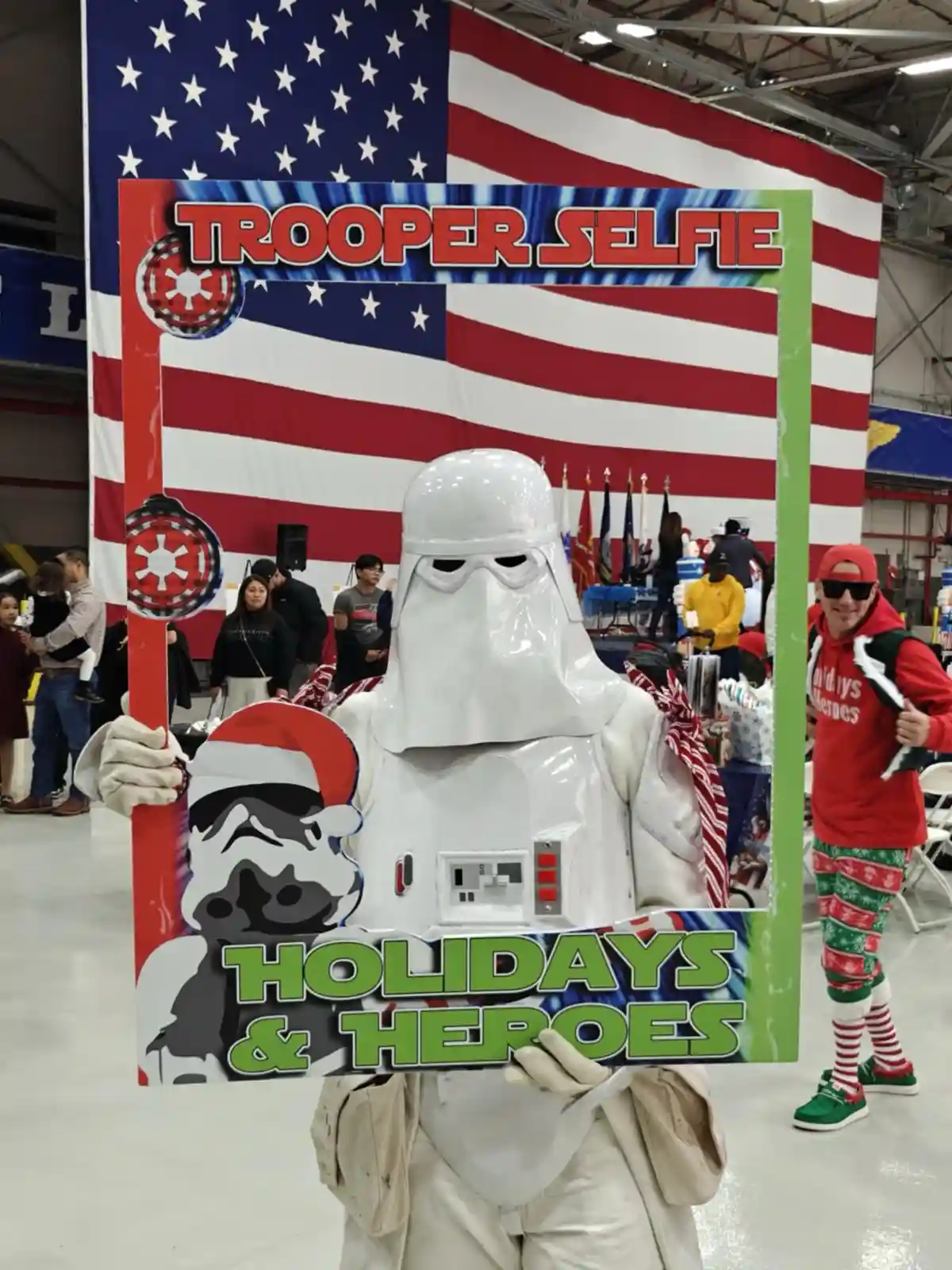 A person in a white Star Wars Stormtrooper costume holds a festive photo frame labeled 'TROOPER SELFIE' and 'HOLIDAYS &…