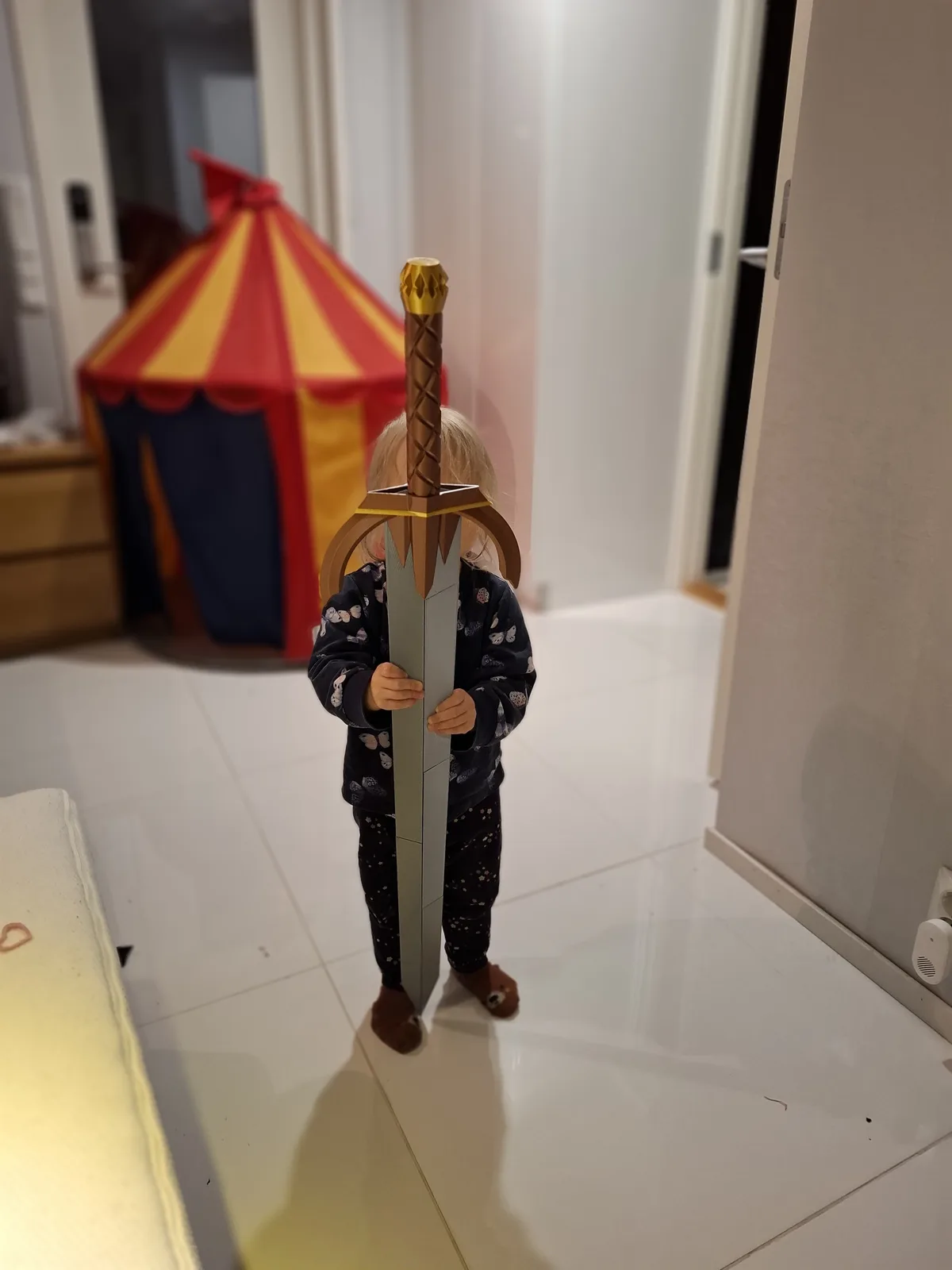 A small child in pajamas stands indoors holding a large, oversized toy sword that completely obscures their face, with a…