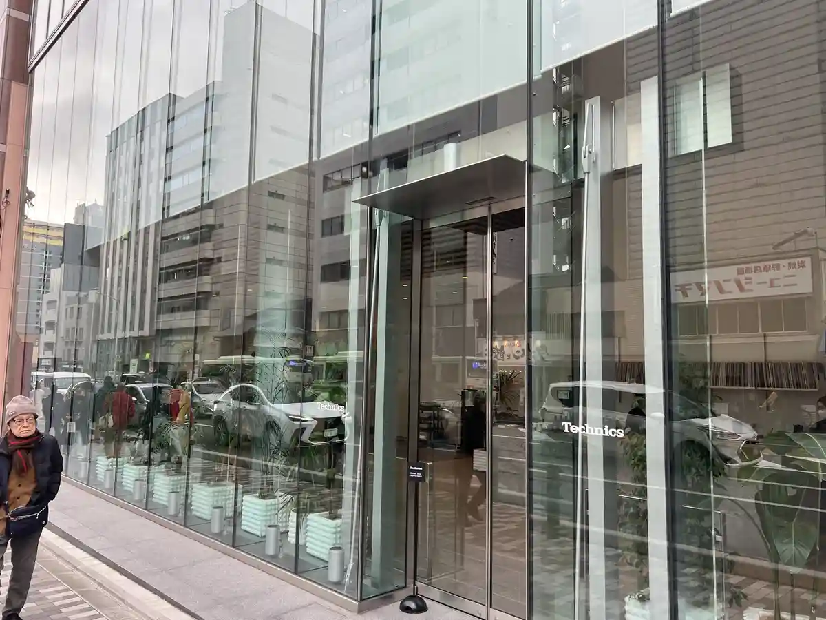 A man in a winter hat and scarf walks past a modern glass-fronted store with 'Technics' branding on the doors. The store's…