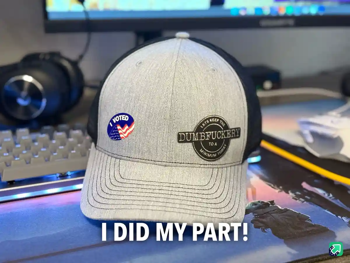 A gray trucker hat sits on a desk with a keyboard and monitor in the background. The hat has two patches: one is a circular…