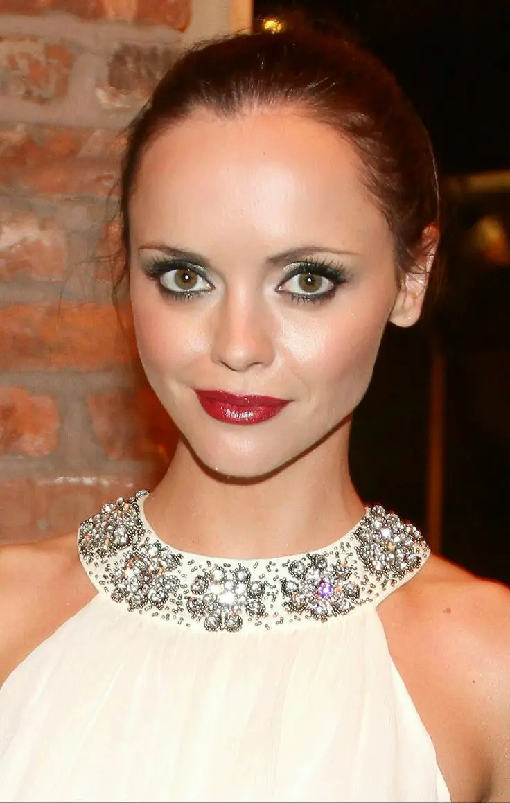 A woman with dark hair pulled back, wearing heavy black eyeliner, glossy red lips, and a white halter top with a jeweled…