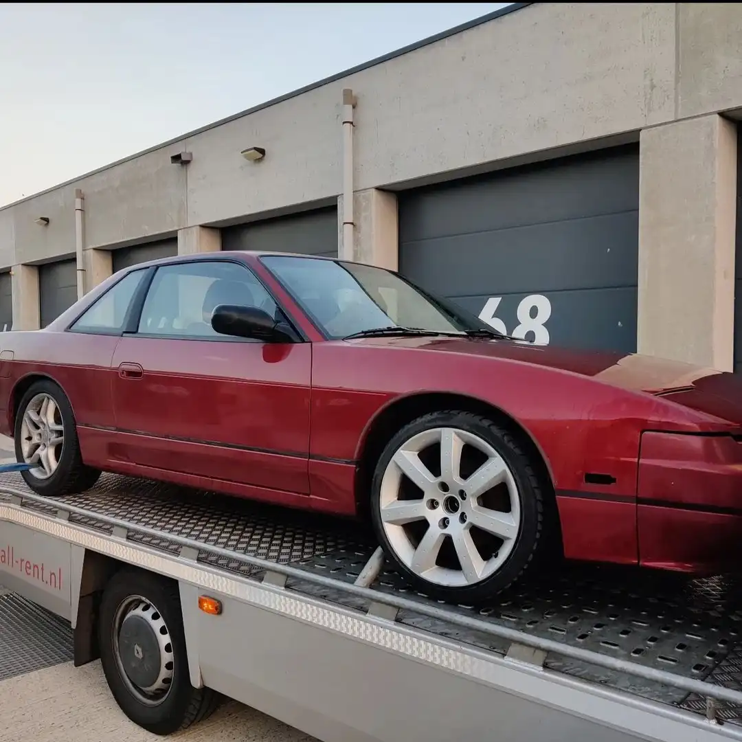 A red Nissan 300ZX sports car is loaded onto a flatbed tow truck in front of a building with garage doors. The car has…