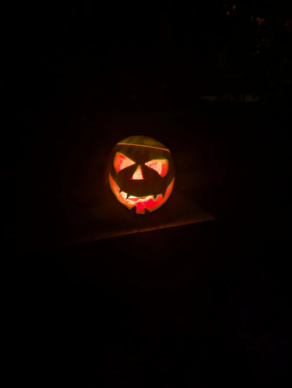 A carved jack-o'-lantern with glowing triangular eyes and a jagged mouth sits on a dark surface at night, illuminated from…