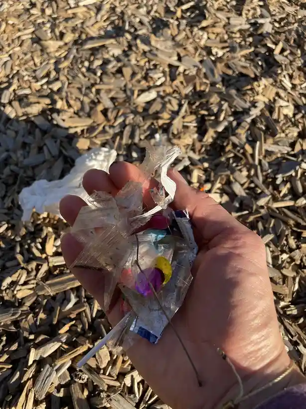 A hand holds a crumpled, dirty plastic bag filled with colorful confetti and a white stick, set against a background of wood…