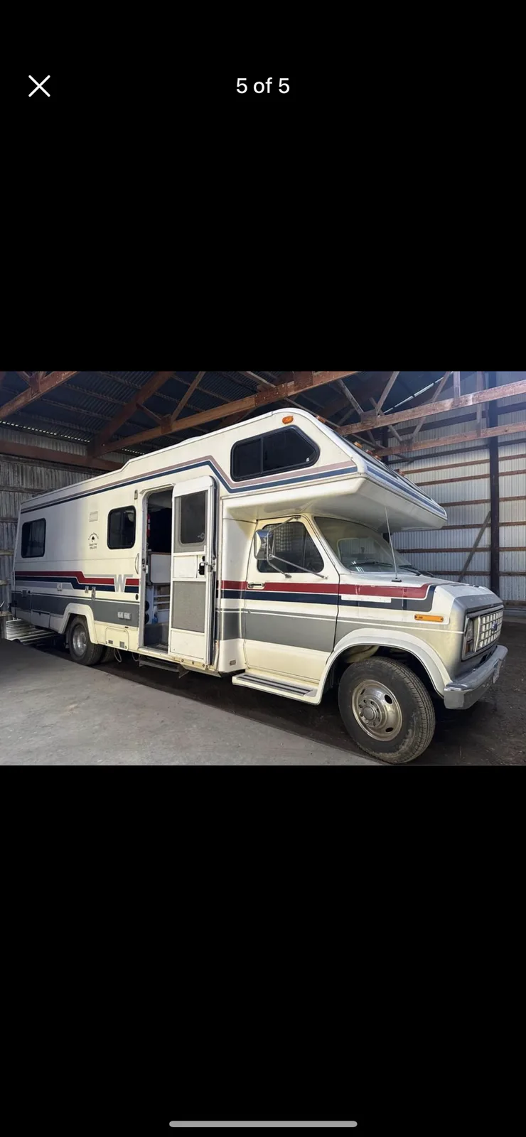 A vintage white, red, and gray motorhome with its side door open is parked inside a metal-roofed warehouse. The overlaid…