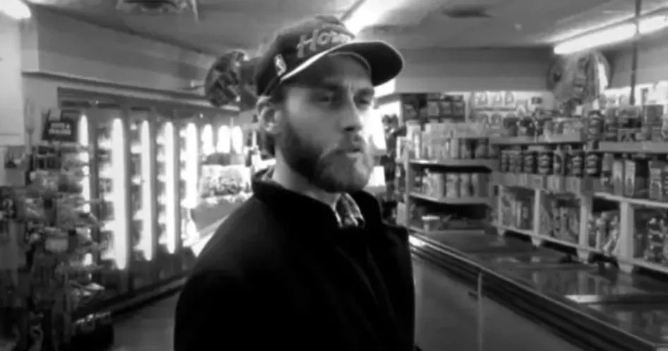 A black-and-white image of a man with a beard and a baseball cap standing in a convenience store aisle, looking annoyed. The…
