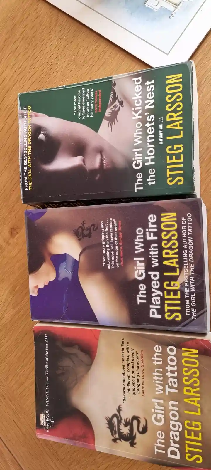 Three physical copies of Stieg Larsson's Millennium series books are stacked on a wooden surface: 'The Girl with the Dragon…
