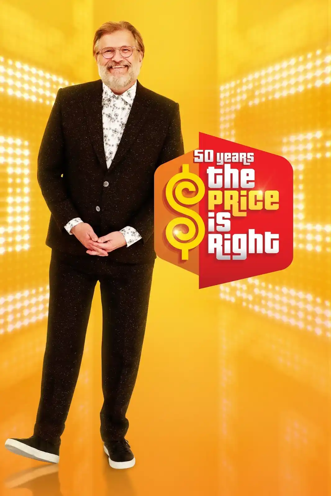 A smiling man with glasses and a beard, wearing a black speckled suit and patterned shirt, stands against a bright yellow…