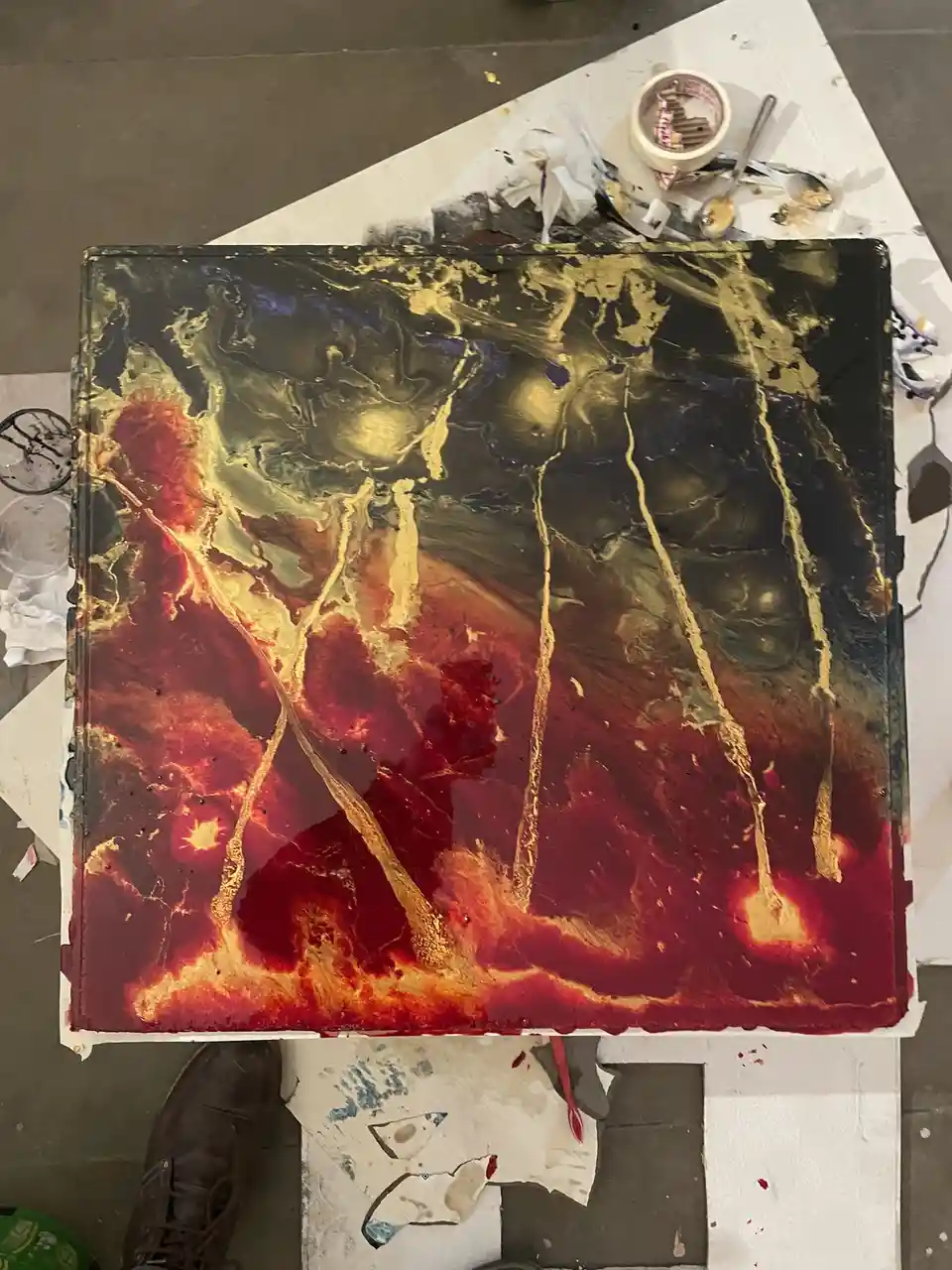 An overhead shot of a large, square, abstract painting with swirling red, gold, and black colors resembling lava and molten…