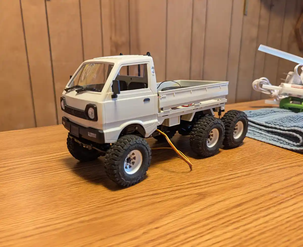 A white remote-controlled truck model with large off-road tires sits on a wooden table, with visible wires extending from…