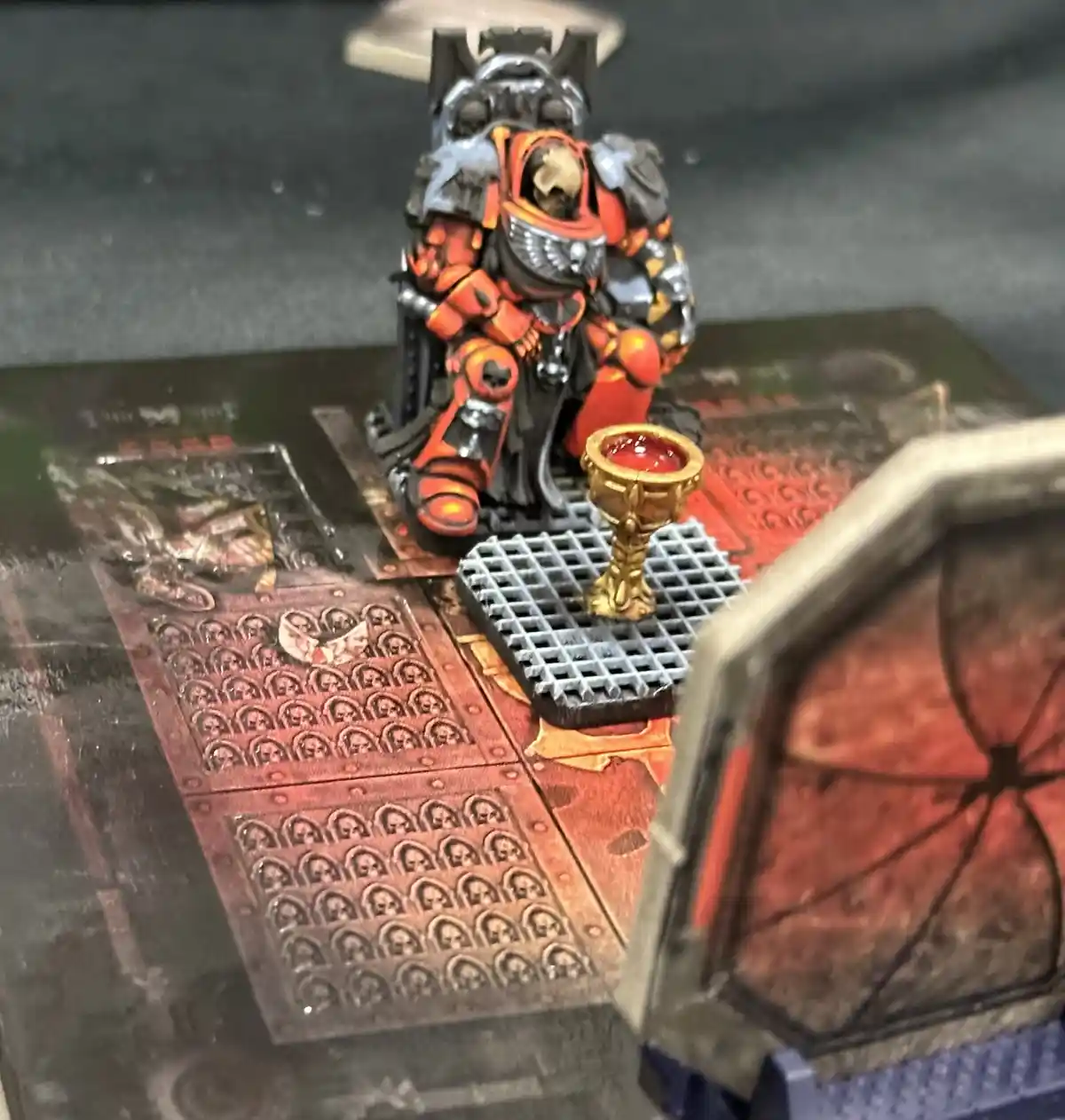 A painted miniature of a heavily armored, orange-and-black space marine sits on a throne-like base next to a golden chalice…
