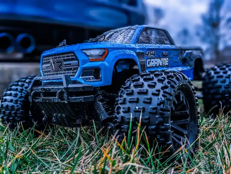 A close-up photo of a blue remote-controlled monster truck with large black tires, covered in water droplets, parked on…