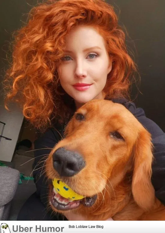 A woman with vibrant red curly hair is smiling while holding a golden retriever that has a yellow tennis ball in its mouth…