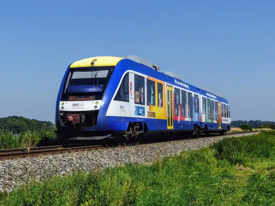 A blue and yellow regional train, branded with 'BRB Bayerische Regiobahn', travels on tracks through a grassy field under a…