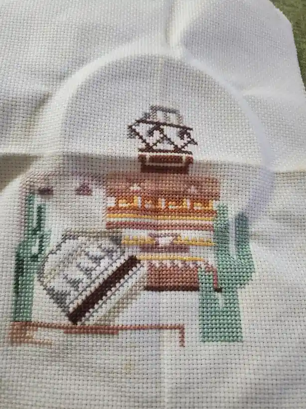 A close-up of a white cross-stitch fabric showing a pixelated, blocky depiction of a desert scene with a saguaro cactus on…