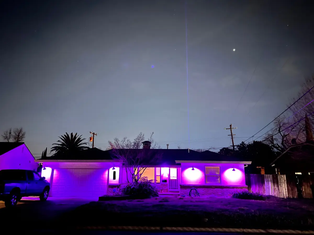 A suburban house at night is bathed in purple light from outdoor fixtures, with a bright vertical laser beam shooting from…