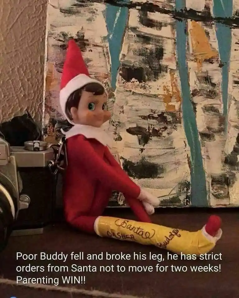 A small red-clothed elf doll, named Buddy, is sitting on a shelf with his right leg in a yellow cast labeled 'Santa's…