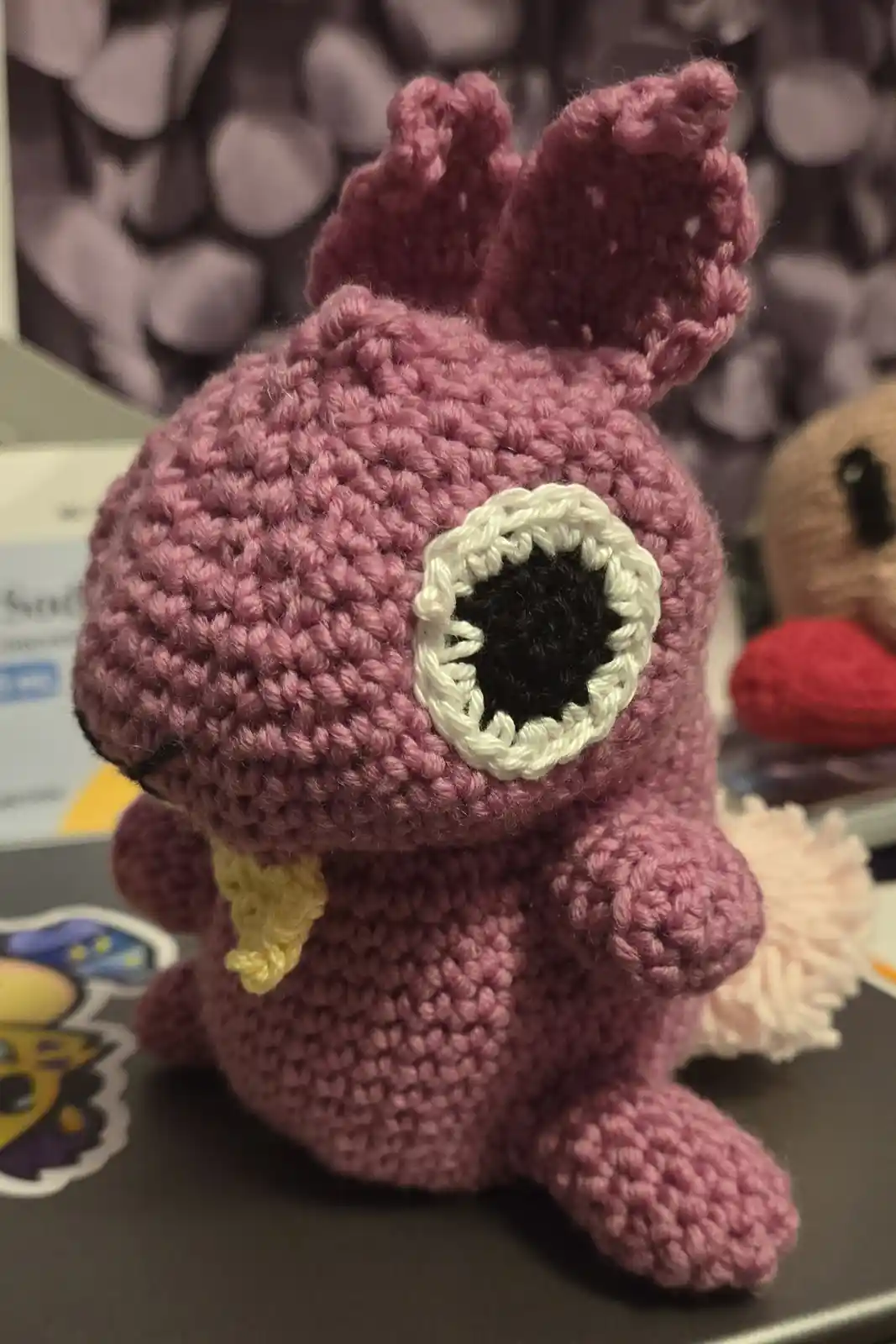 A handmade pink crocheted plush toy resembling a small, cute creature with large, round eyes made of black and white yarn…