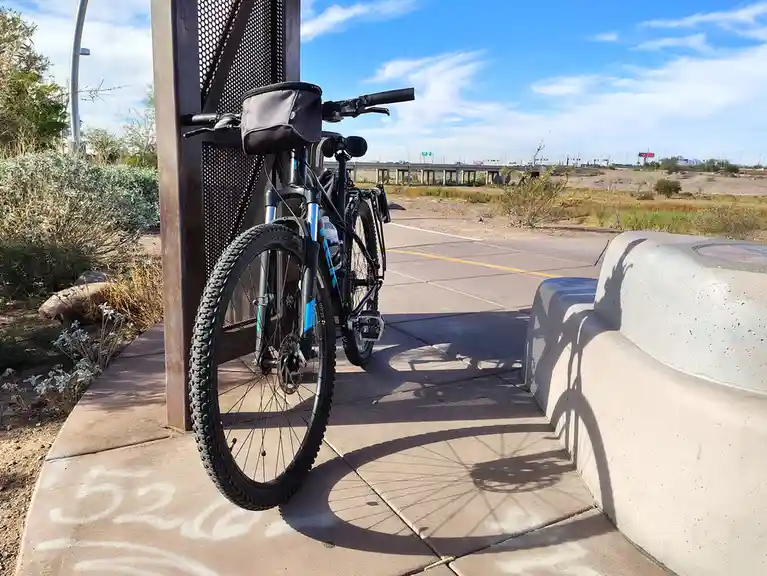 A mountain bike is parked next to a metal post on a paved path in a dry, arid landscape under a bright blue sky. The bike's…