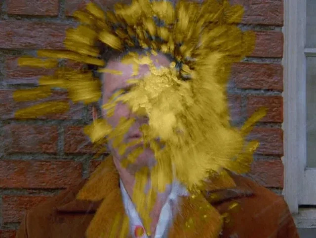 a man in a brown coat stands against a brick wall, his face and hair obscured by a yellow paint explosion effect. The…