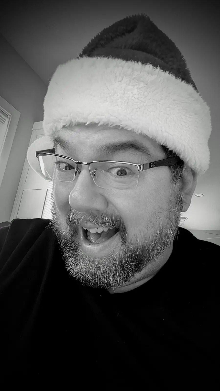 A black-and-white selfie shows a man with glasses and a beard wearing a Santa hat, making a wide-eyed, open-mouthed…
