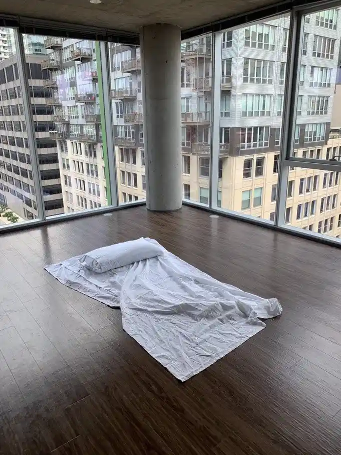 A minimalist apartment with dark wood floors and floor-to-ceiling windows overlooking a dense urban skyline. A single white…