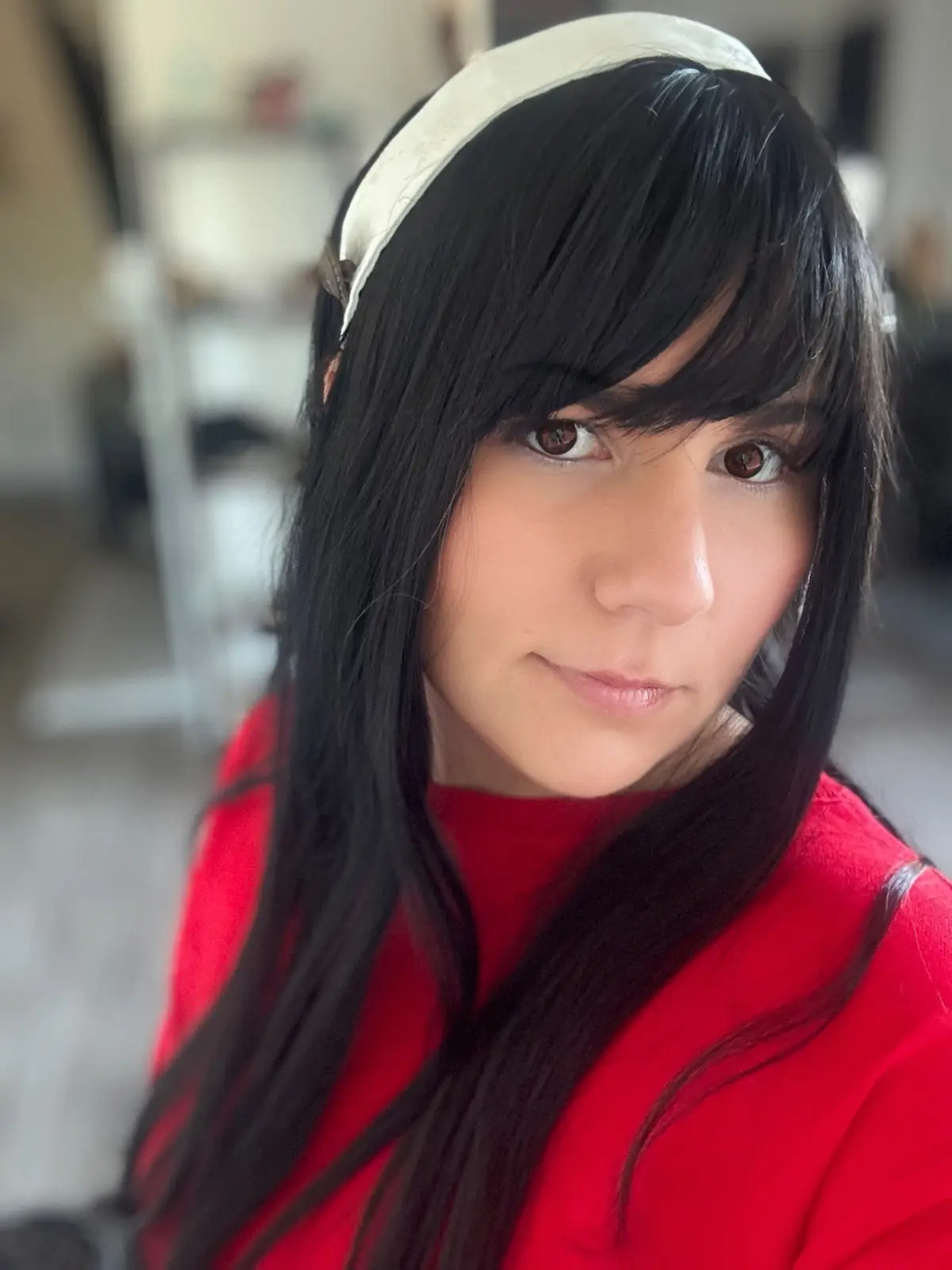 A person with long black hair and a white headband wears a red top and stares directly at the camera with a subtle, slightly…