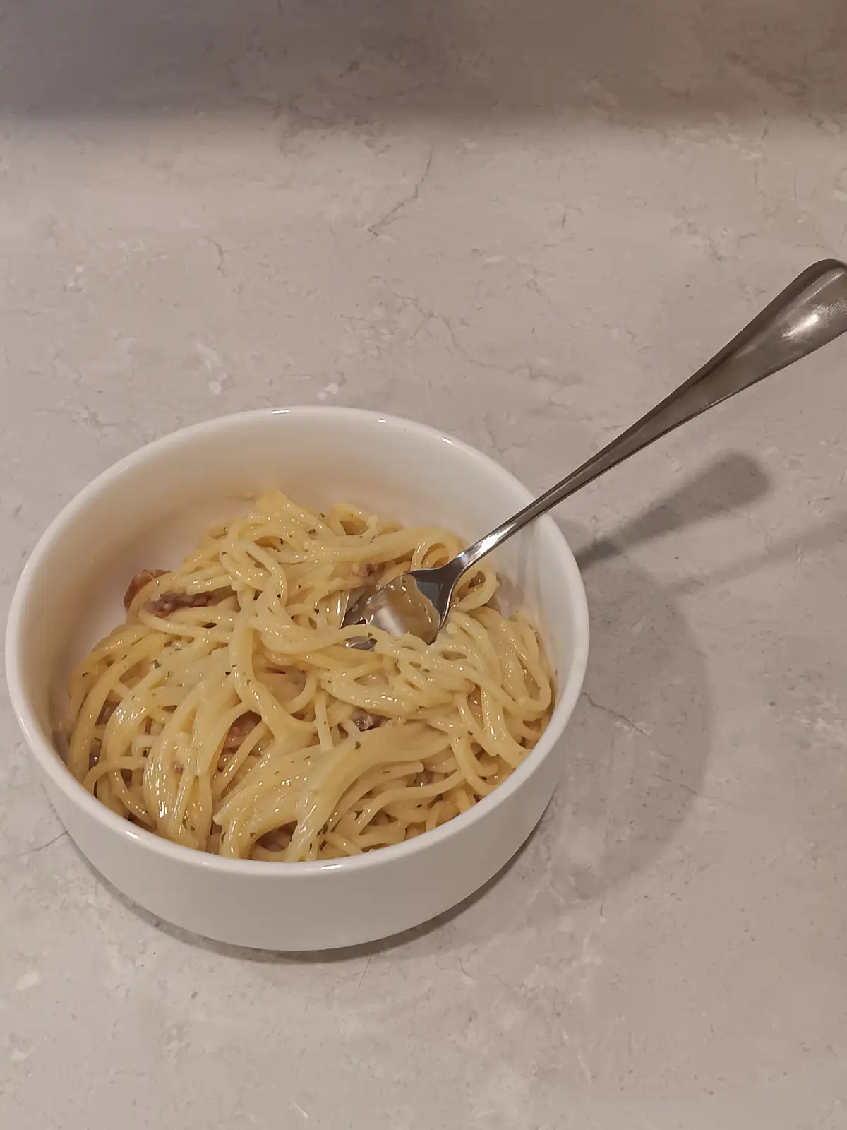 A white bowl filled with spaghetti and small pieces of meat sits on a light gray marble countertop, with a metal fork…