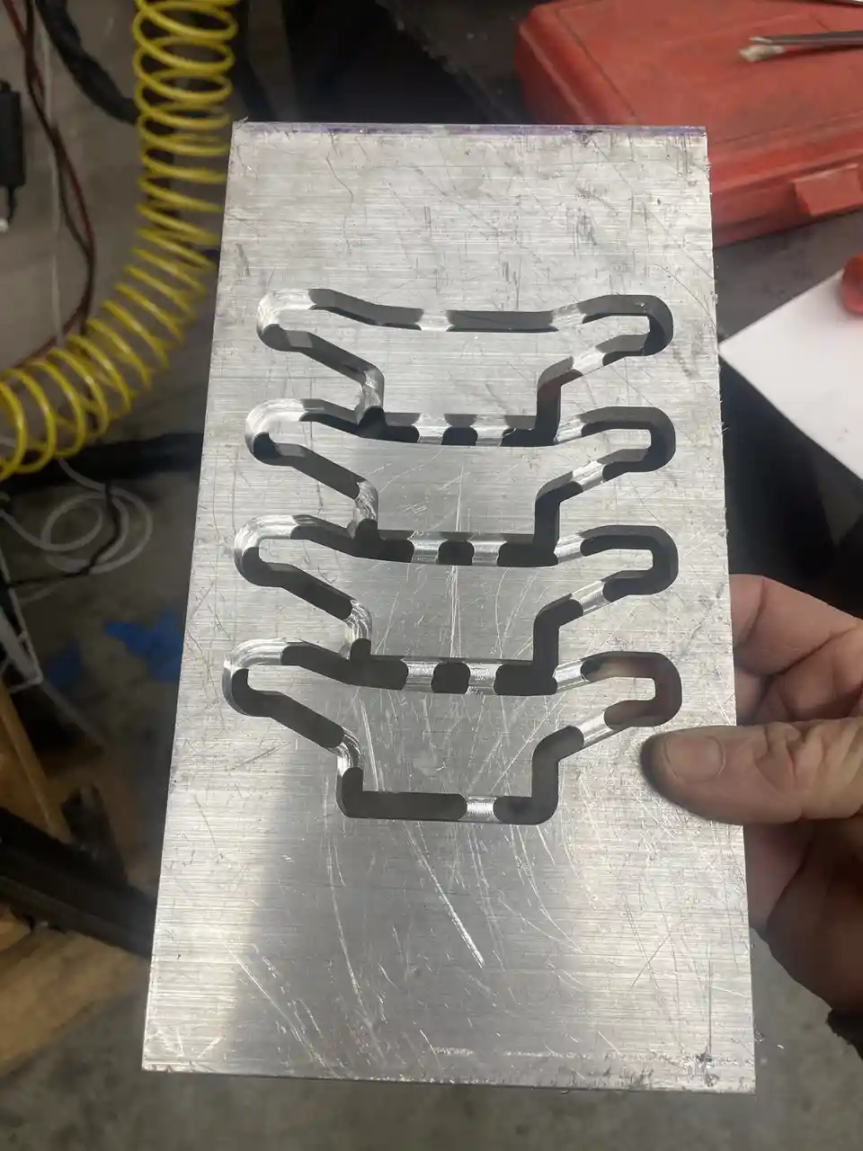 A hand holds a brushed metal sheet with four identical cutouts shaped like a race track circuit, each with multiple turns…