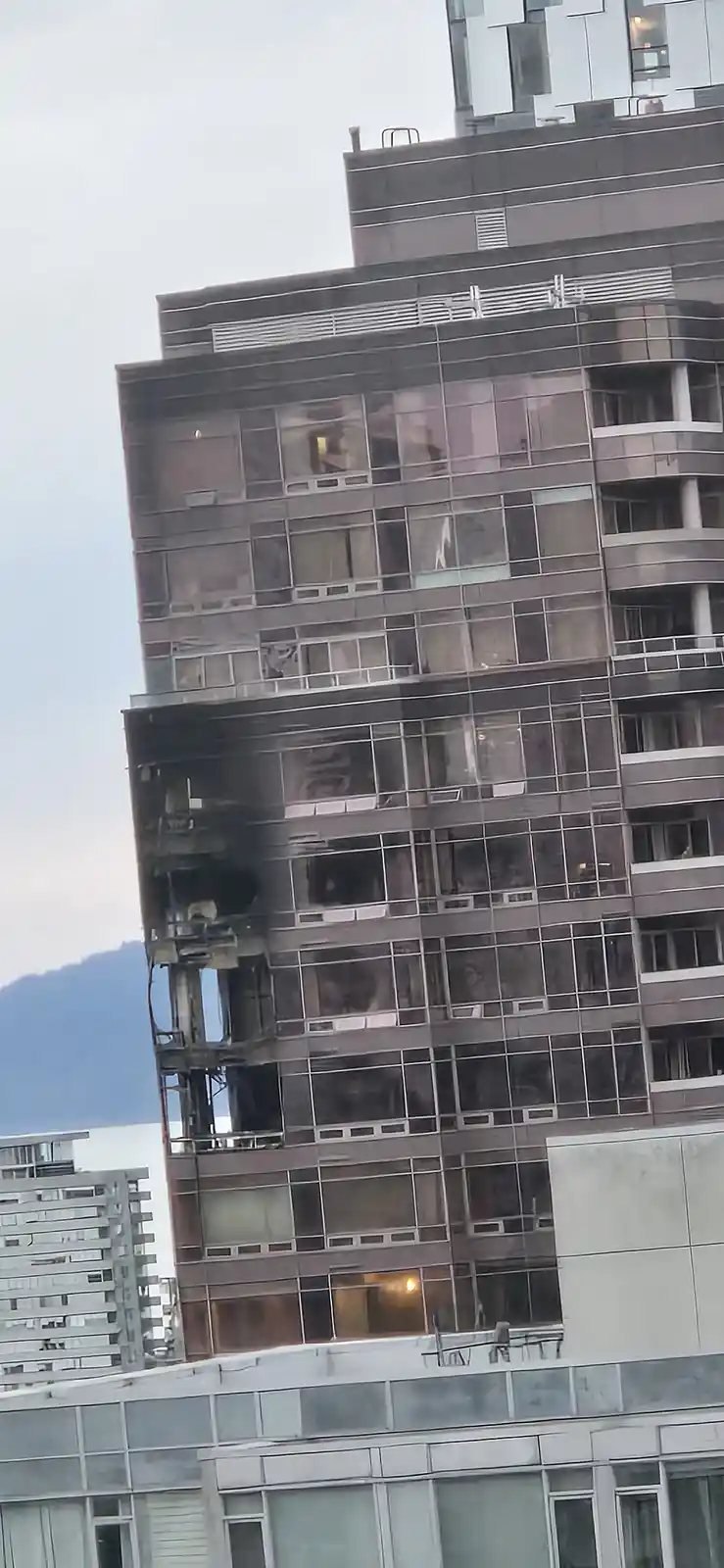 A tall modern apartment building shows visible damage from a fire, with charred exterior walls, broken windows, and smoke…