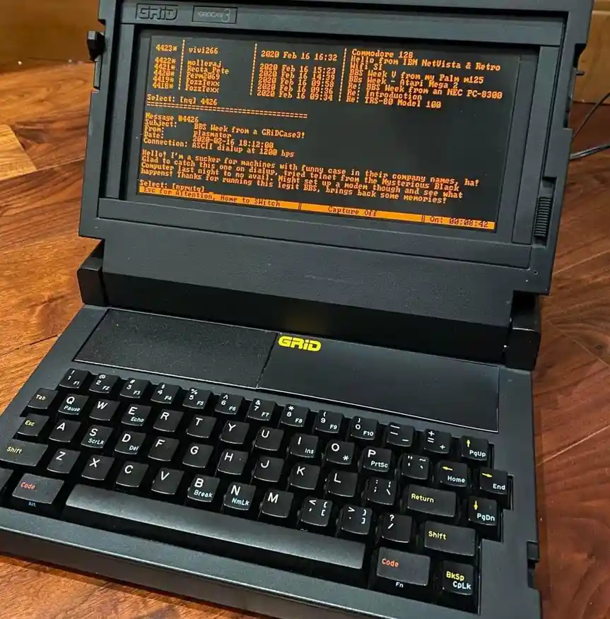 A black, retro-style laptop with 'GRID' branding on the keyboard and screen bezel, displaying a BBS (Bulletin Board System)…