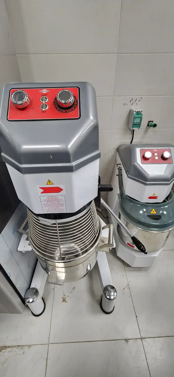 Two large industrial dough mixers sit side by side in a tiled kitchen, one larger with a red control panel and the other…