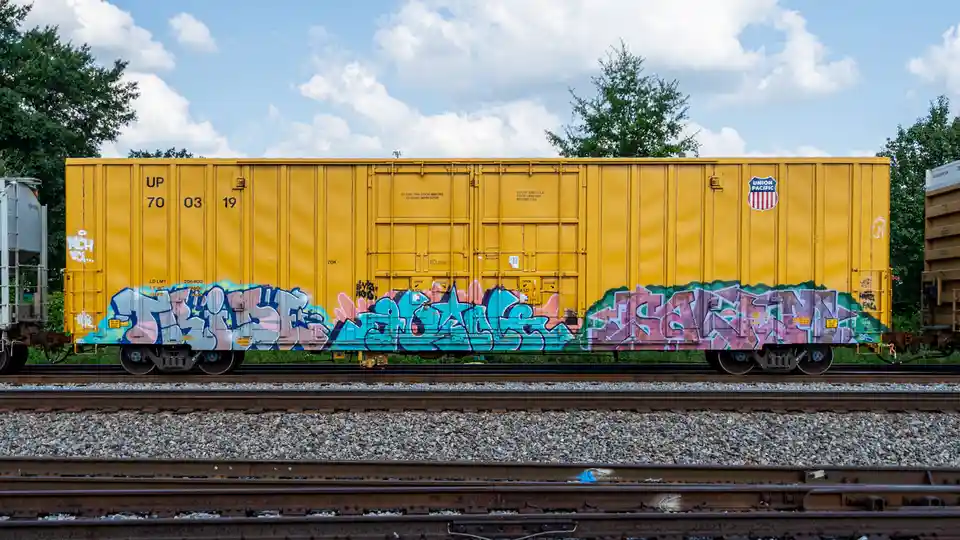 A yellow Union Pacific freight train car covered in colorful graffiti art sits on railway tracks under a partly cloudy sky…