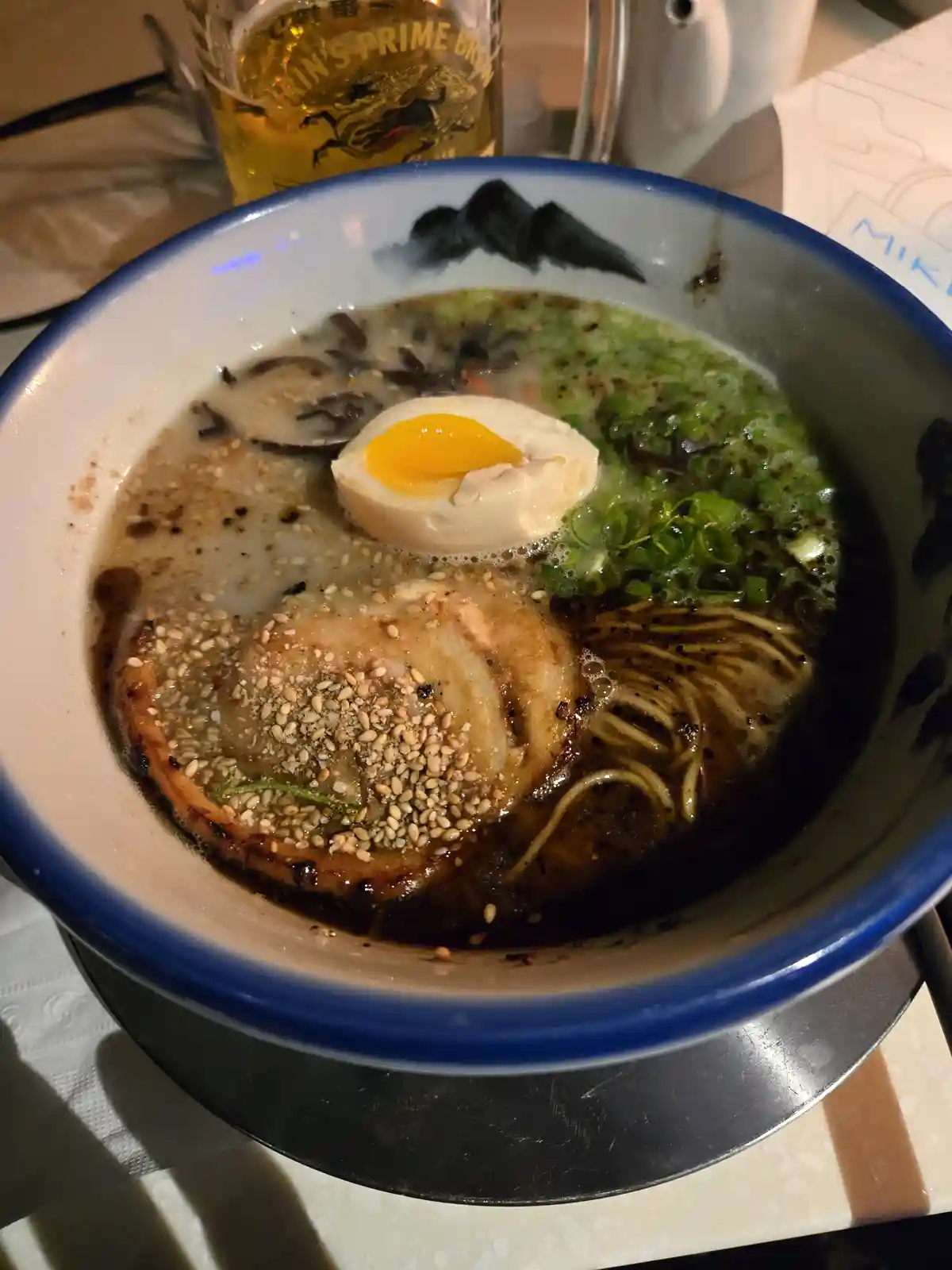 a close-up photo of a steaming bowl of ramen with a soft-boiled egg, chashu pork, green onions, and sesame seeds in a dark…