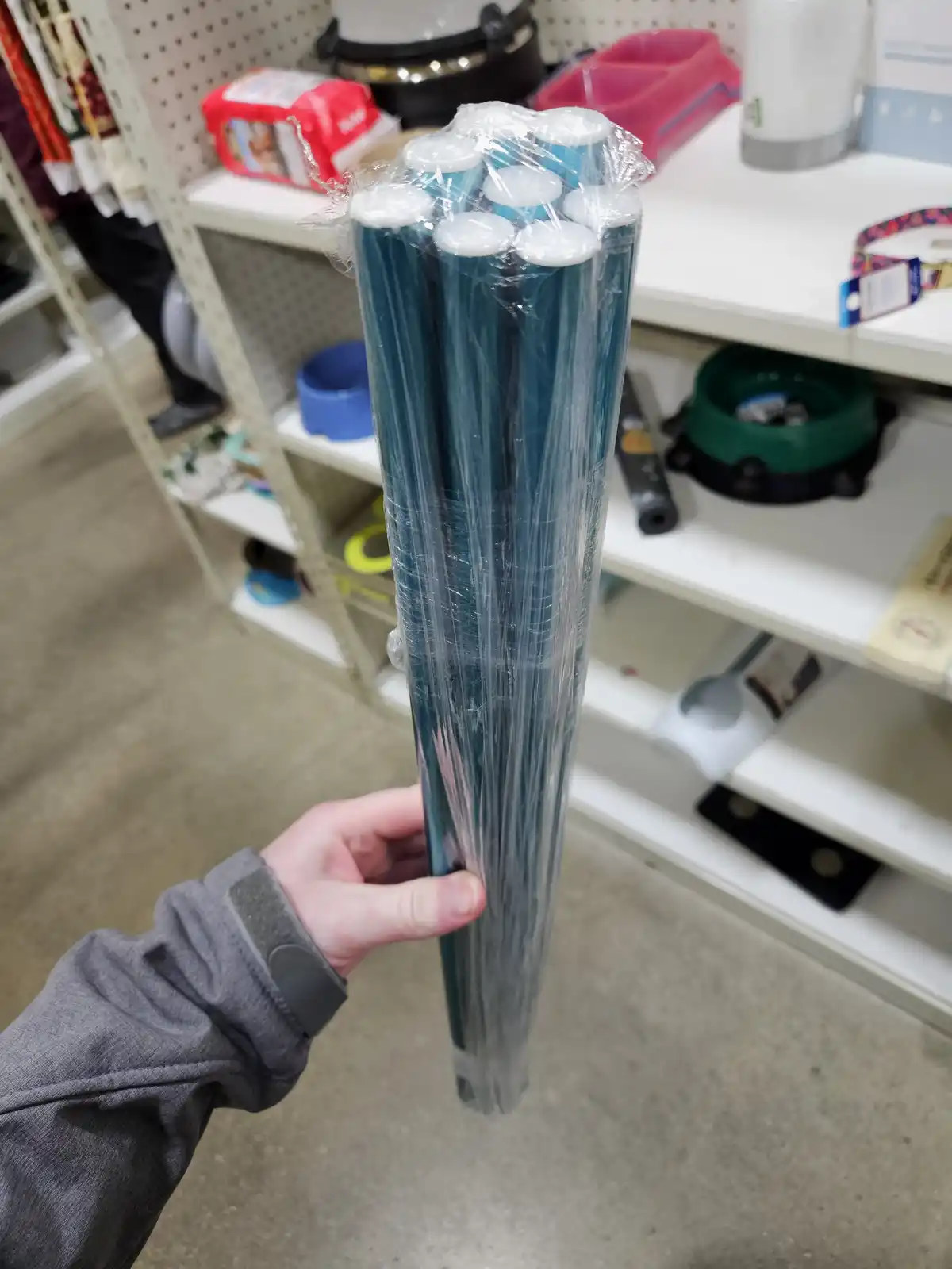 A person's hand holds a large bundle of teal-colored plastic sticks, wrapped in clear plastic, in what appears to be a…