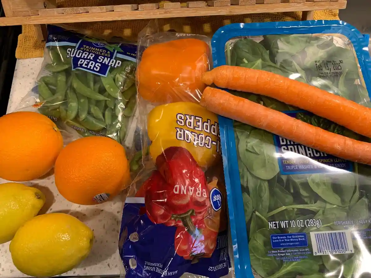 A grocery store produce display featuring sugar snap peas, orange and yellow bell peppers, carrots, spinach, oranges, and…