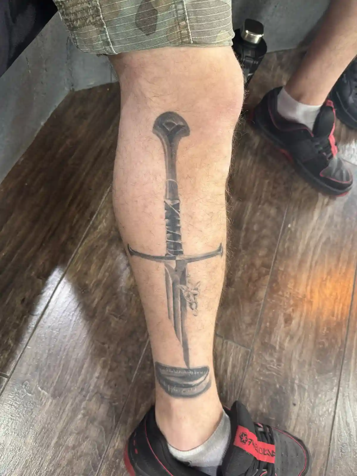 A close-up photo shows a person's lower leg with a detailed black-and-gray tattoo of a sword running vertically from above…