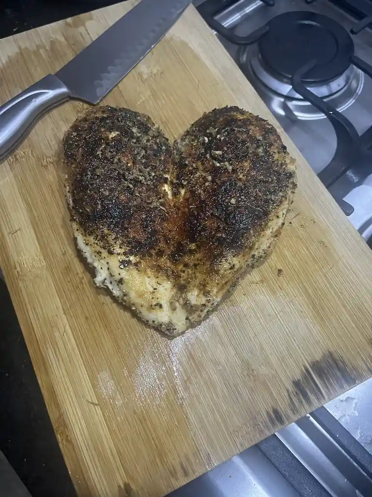 A cooked, heart-shaped chicken breast sits on a wooden cutting board next to a knife, with a stovetop visible in the…