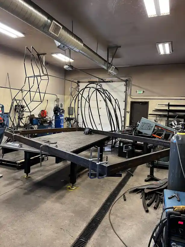A metal fabrication workshop with welding equipment, a large metal workbench, and two large metal sculptures in progress…
