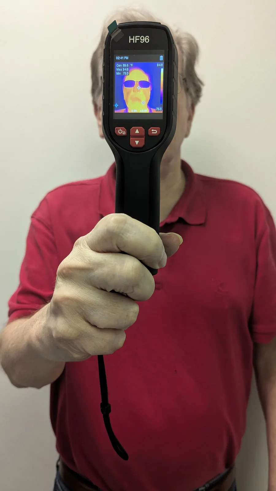 A man in a red polo shirt holds a thermal imaging camera, which displays a thermal scan of his own face on its screen. The…