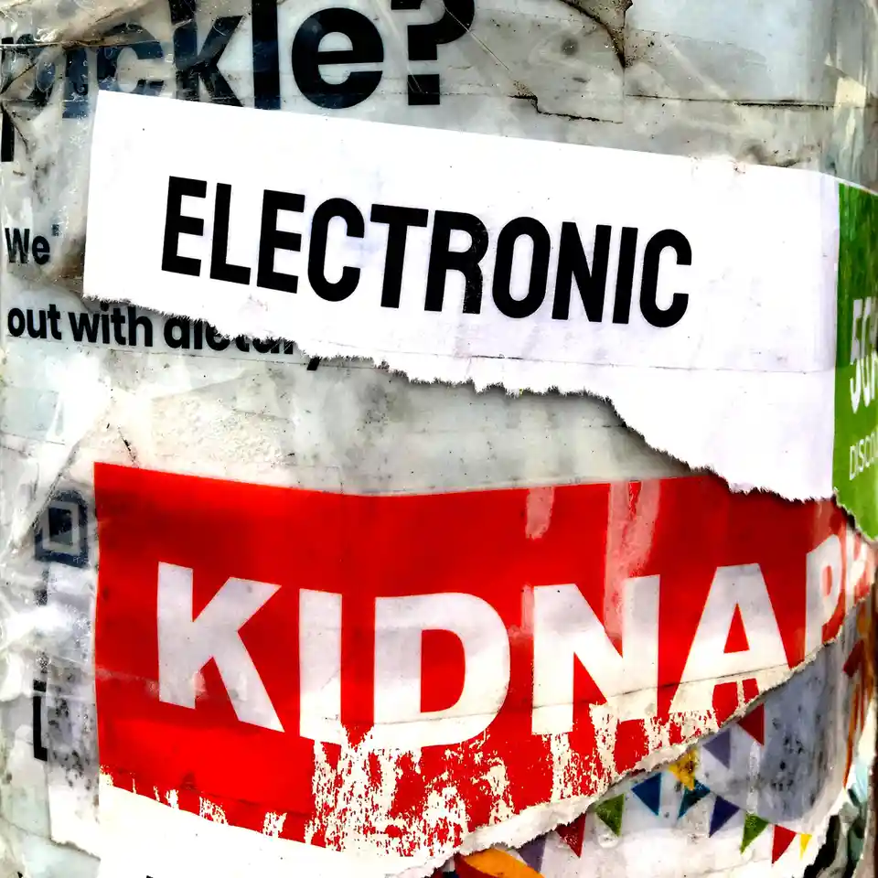 A close-up of a weathered, layered wall covered in torn posters, with a prominent white sticker reading 'ELECTRONIC'…