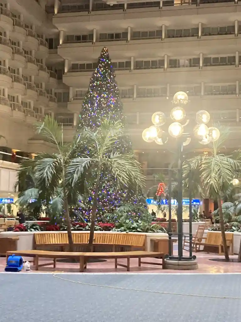 A large, brightly lit artificial Christmas tree stands in the center of a spacious, multi-level indoor atrium, surrounded by…
