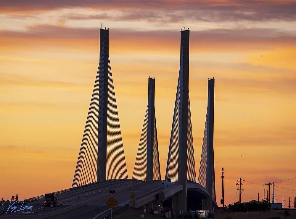 A photograph of a cable-stayed bridge at sunset, with four tall pylons and orange traffic cones lining the road. The sky is…