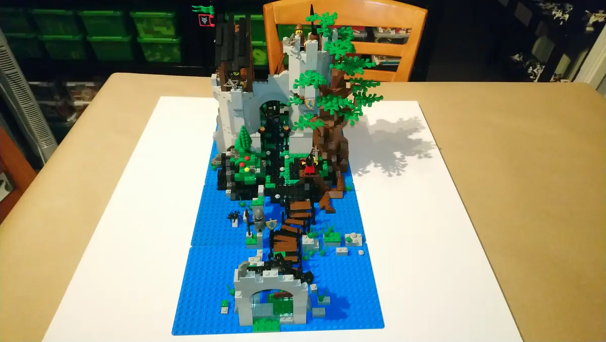 A detailed LEGO castle built on a blue baseplate representing water, featuring a large tree with green foliage, a stone…